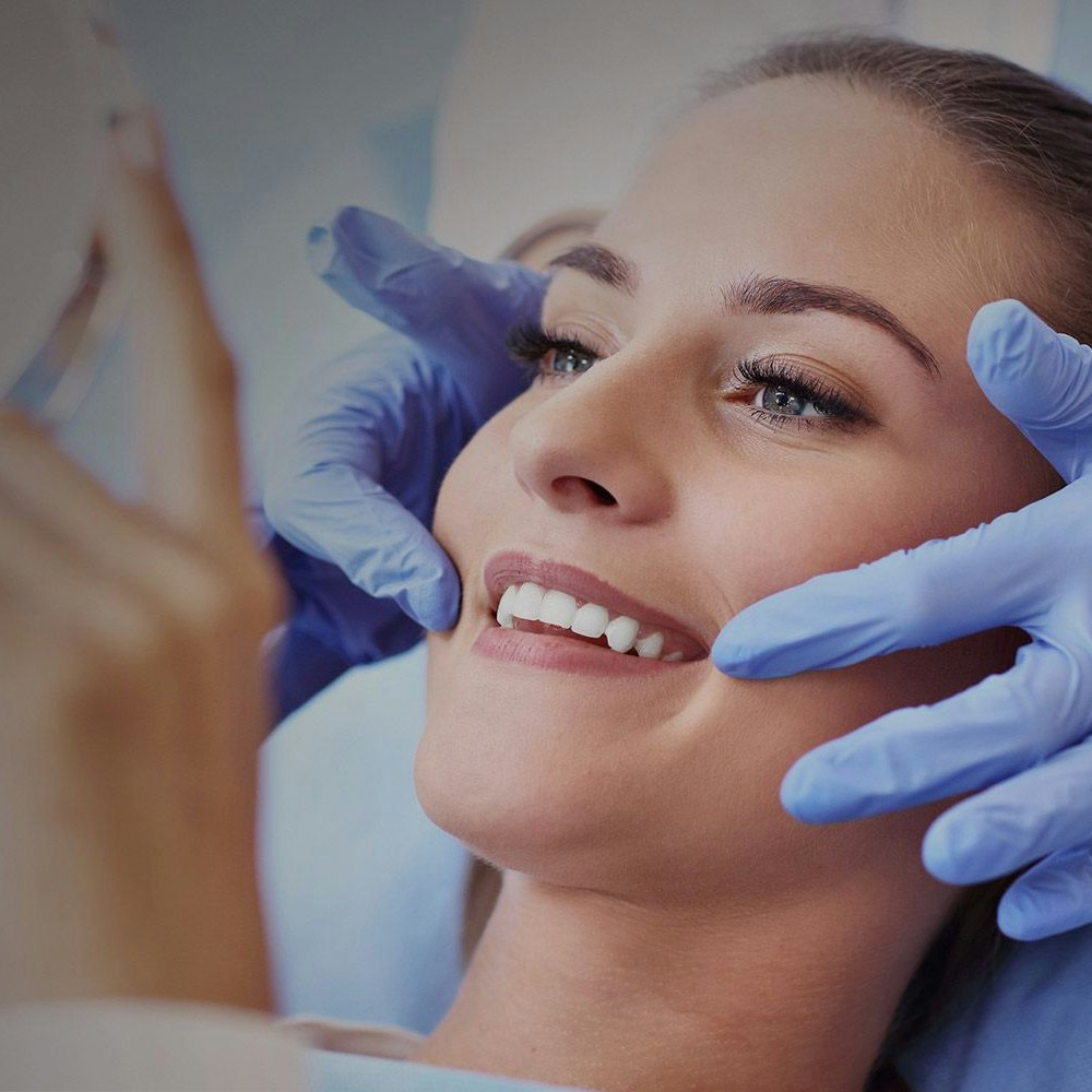 Woman looking at her smile in the mirror while the dentist's gloved hands hold her head